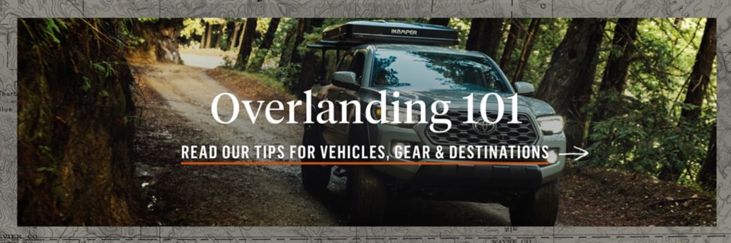 A truck drives down a forested dirt road. Text overlay reads: Overlanding 101, read out tips for vehicles, gear & destinations.